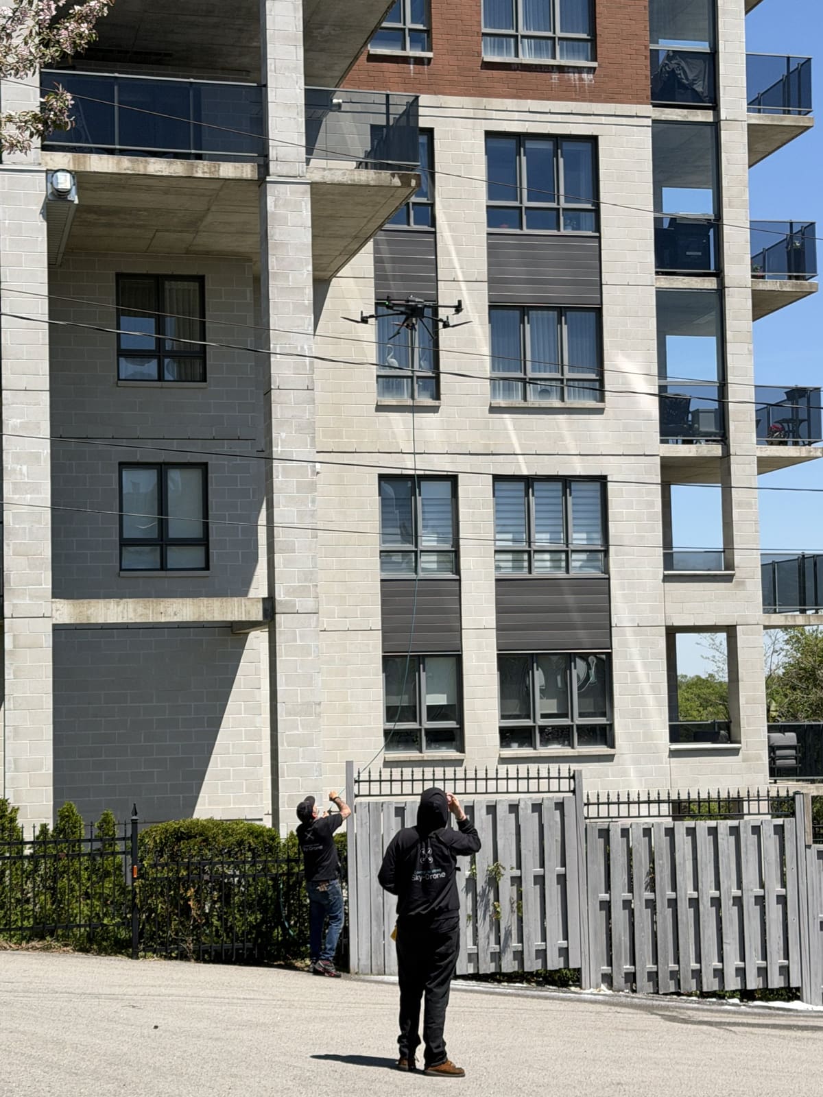 Deux opérateurs au sol, au pied d'une façade de condos, le drone au travail devant une fenêtre au-dessus d'eux.